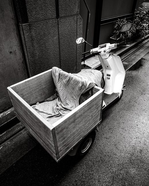 A black and white image showing a small delivery scooter parked next to a building. Attached to the scooter is a large, rectangular wooden crate lined with fabric, suggesting it is a flower delivery v