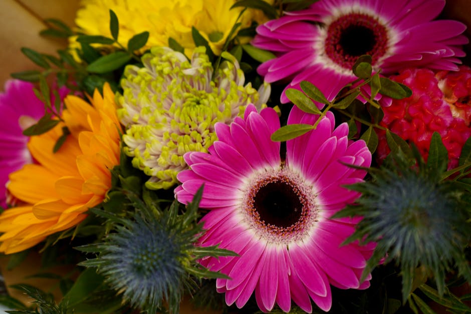 A vibrant floral arrangement featuring a mix of fresh flowers including large pink gerbera daisies with dark centers, bright yellow sunflowers with layered petals, delicate green and yellow chrysanthe