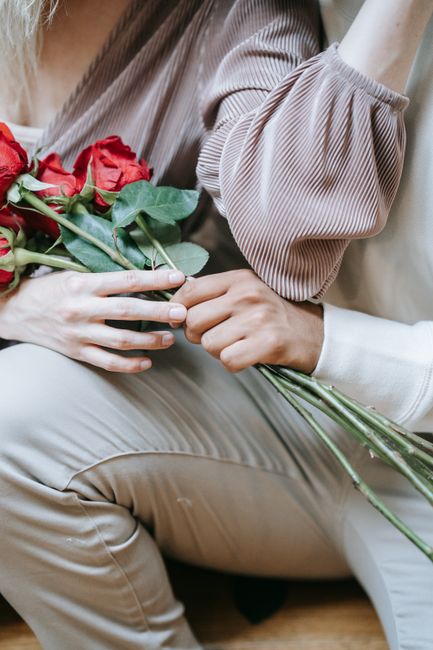 A close-up of two individuals exchanging a floral gift, one person holding a bouquet of fresh red roses with green stems and leaves, wrapped in simple transparent material. The recipient's hand is rea