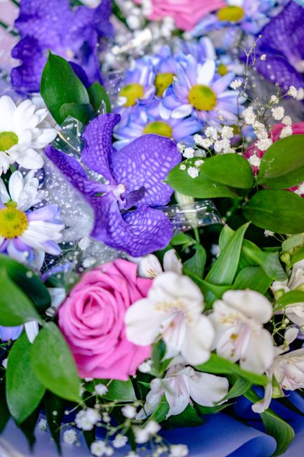 A vibrant floral arrangement featuring a variety of fresh flowers including purple irises with textured petals, white daisies with yellow centers, soft pink roses, and delicate white orchids, all comp
