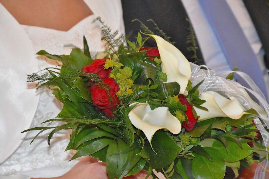 A floral arrangement featuring deep red roses and elegant white calla lilies, complemented by lush green foliage and delicate greenery accents. The bouquet is wrapped in clear cellophane with a decora